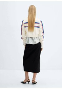 Zkrácená krémová bunda s modrými a červenými pruhy, vrstvená přes skládanou pruhovanou košili, spárovaná s černou midi sukni a černými botami.