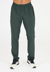 Donkergroene sportbroek voor heren met een trekkoord in de taille, zijzakken met rits en een gladde, lichte stof. Gecombineerd met zwart-witte sneakers.