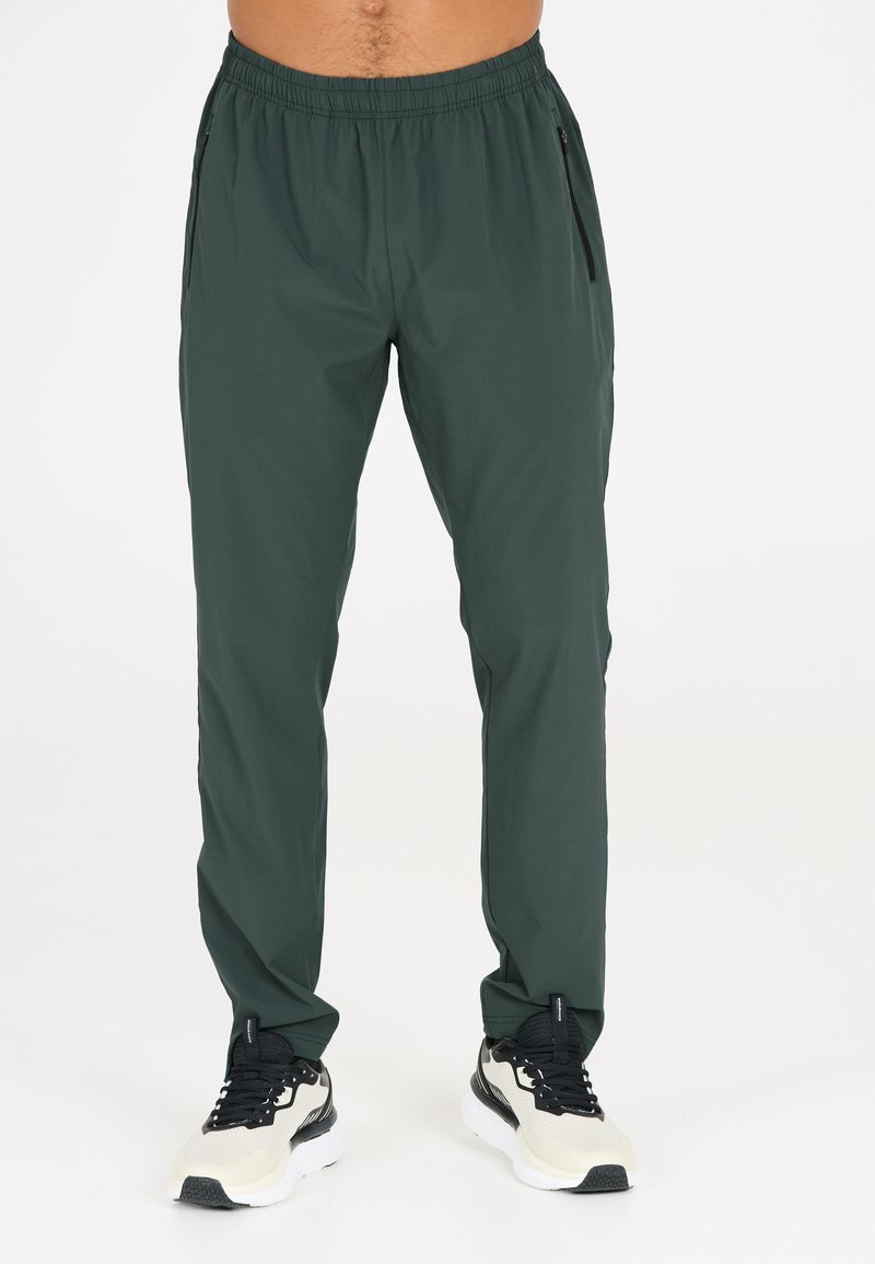 Donkergroene sportbroek voor heren met een trekkoord in de taille, zijzakken met rits en een gladde, lichte stof. Gecombineerd met zwart-witte sneakers.