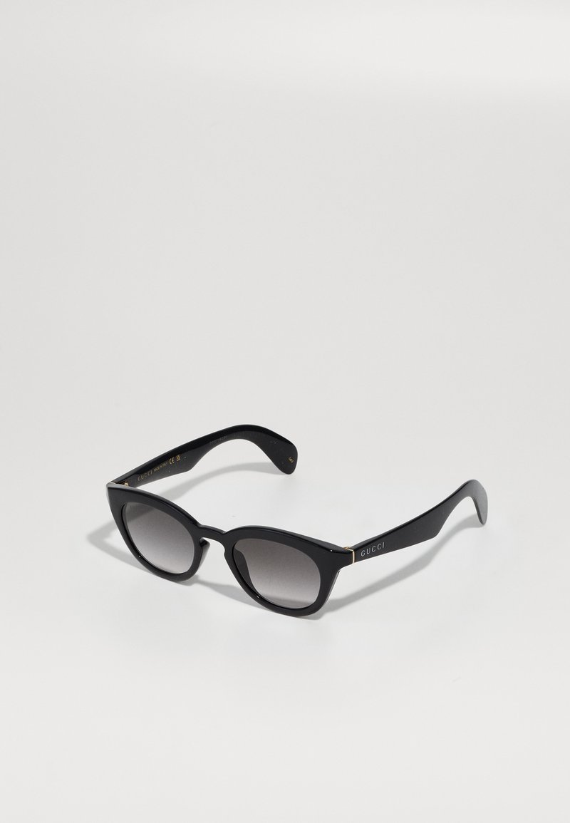 Ochelari de soare Gucci, culoare neagră cu formă de ochi de pisică, cu lentile gri degradate, brațe late și numele brandului vizibil pe tâmplă, așezați pe o suprafa�ță albă.