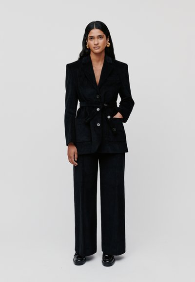 Blazer en corduroy noir avec une taille ceinturée, de grands boutons et des poches avant, associé à un pantalon large en corduroy noir.