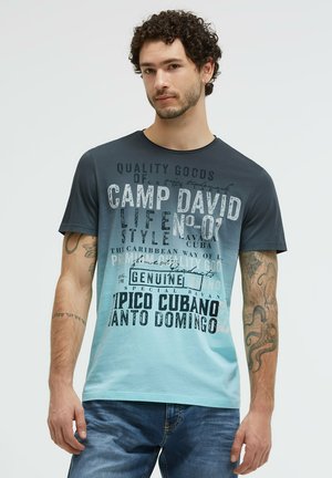Junger Mann mit lockigem Haar, der ein blaues Gradient Camp David T-Shirt mit großem Aufdruck und blaue Jeans trägt und vor einem einfachen Hintergrund steht.