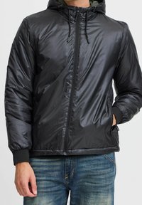 Schwarze, leichte Steppjacke mit Kapuze, Reißverschluss, gerippten Bündchen und Seitentaschen. Glatte Textur und glänzendes Finish.