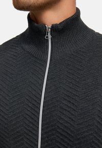 Suéter negro con cremallera y cuello alto, que presenta un patrón de espiga texturizado y detalles de cremallera plateados en el frente.