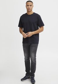 Tricou negru cu guler rotund, cu mâneci scurte, asortat cu blugi slim-fit gri și adidași închiși la culoare, pe un fundal neutru.
