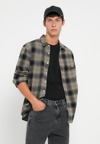 Camisa de cuadros abotonada en tonos beige, negro y gris; llevada sobre una camiseta negra. Combinada con jeans en un gris oscuro, de diseño minimalista. Gorra negra.