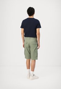 T-shirt in cotone blu navy e pantaloni cargo verde oliva con multiple tasche. Calze bianche e sneakers di colore chiaro completano l'outfit.