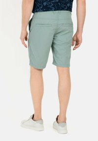 Hellgrüne Shorts mit glatter Textur, zwei Gesäßtaschen und umgeschlagenen Säumen, kombiniert mit weißen Sneakern und einem gemusterten dunkelblauen Hemd.