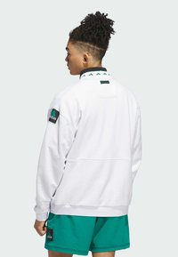 Felpa bianca con dettaglio verde sul colletto, toppa con logo sul lato e vestibilità ampia. Abbinata a pantaloni shorts turchesi, che mostrano uno stile sportivo e informale.