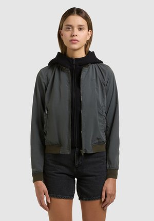 Joven mujer de pie mirando hacia adelante, con una chaqueta bomber gris sobre una sudadera con capucha negra y pantalones cortos de mezclilla negros, sobre un fondo liso.