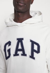Felpa color crema con un grande logo "GAP" navy sul davanti. Tessuto morbido e strutturato con cappuccio regolabile e vestibilità comoda.