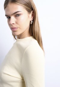 Νεαρή γυναίκα με ίσια ανοιχτό καστανά μαλλιά, ντυμένη με κρεμ μακρυμάνικη μπλούζα με ριγέ υφή και ασημένια σκουλαρίκια κρίκους, κοιτάζοντας ελαφρώς προς το πλάι.