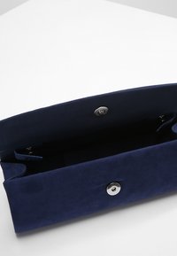 Pochette en daim bleu marine avec une texture lisse, dotée d'une fermeture à bouton-pression en argent et d'un intérieur spacieux. Aucun poche extérieure visible.