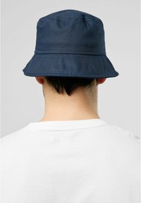 Mörkblå bucket-hatt med en bred, utbuktande skärm, tillverkad av textil med struktur, med synliga sömnadsdetaljer, sedd bakifrån.