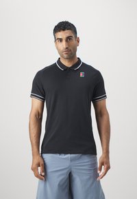 Svart polo-skjorte med hvite aksentstriper på kragen og ermene. Har en liten fargerik logo på venstre bryst.