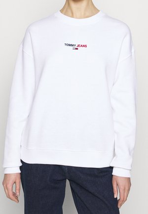 Biała bluza z napisem "Tommy Jeans" i logo umieszczonym na środku na klatce piersiowej, noszona z ciemnoniebieskimi dżinsami na stojącej osobie.