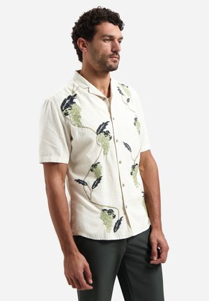 Mann trägt ein cremefarbenes Kurzarmhemd mit bestickten grünen Traubenclustern und schwarzen Blättern, kombiniert mit dunkler Hose, steht seitlich.