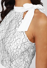 Robe blanche avec un dessus en dentelle florale noire, présentant un col haut et un détail de nœud sur le côté de l'épaule. Texture lisse.