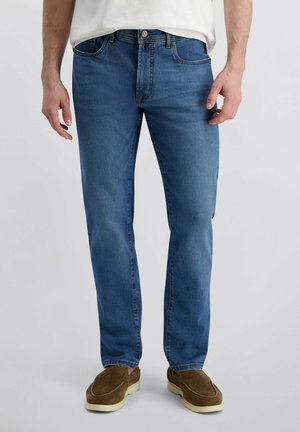 Mann mit gerade geschnittenen blauen Jeans, weißem Hemd und braunen Loafers steht vor einem schlichten hellen Hintergrund.