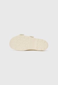 Lys beige sandal såle med et teksturert grep mønster og to justerbare stropper på oversiden. Laget av et slitesterkt, fleksibelt materiale.