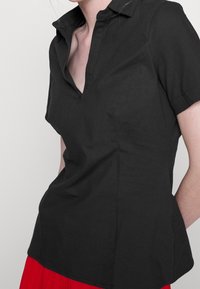 Camicia nera a maniche corte con colletto a punte, caratterizzata da una vestibilità strutturata e una texture liscia, completata da un leggero spacco laterale.