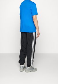 Blå bomullströja, svarta träningsbyxor med vita sidostripes och ljusgrå sneakers. Enkla, avslappnade träningskläder från baksidan.