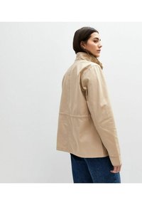 Beige katoenen jack gezien vanaf de achterkant, met een opstaande kraag, casual pasvorm, zijnaad en een effen textuur. Draagt over blauwe denim.