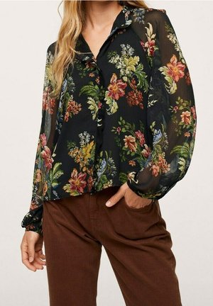 Blusa negra con estampado floral y tela transparente, con patrones de flores coloridas, botón al frente y mangas abullonadas; combinada con pantalones marrones.