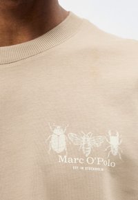 Közelkép bézs ingről, világos színű rovar illusztrációkkal és a mellkason található „Marc O'Polo EST. IN STOCKHOLM“ felirattal.