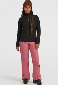 Zwart zip-neck top en roze sneeuwbroek. De broek heeft een gestructureerde afwerking, terwijl de top lange mouwen en een slanke pasvorm heeft. Zwarte skiboots.