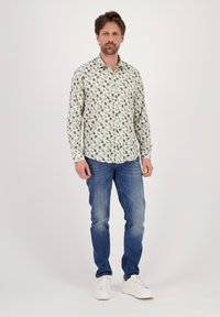 Camicia bianca con bottoni caratterizzata da un motivo floreale verde, abbinata a jeans blu e sneaker bianche. La camicia ha un colletto classico e maniche lunghe.