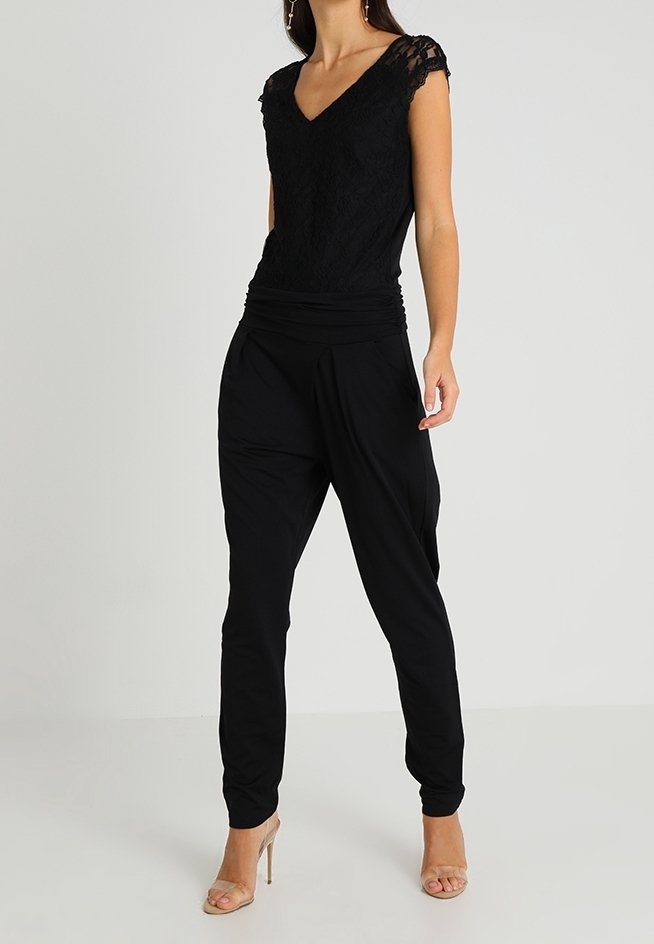 Femme portant un top noir en dentelle à manches courtes et un pantalon noir ample avec des sandales beiges à talons hauts, debout devant un fond uni.
