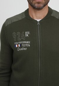 Olivovo zelený sveter na zips s pletenou textúrou, ktorý má tmavé ramenné panely a vyšívaný text na prednej strane.