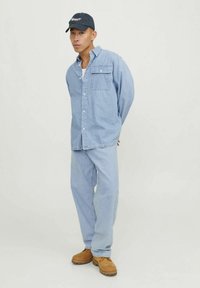 Svetlo modra denim srajca z žepom na prsih, v kombinaciji z ujemajočimi svetlo modrimi hlačami in rjavimi čevlji. Črna kapa z belo logotipom.