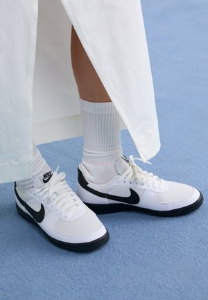 Osoba nosząca białe buty sportowe Nike z czarnym logo i białe prążkowane skarpetki za kostkę, stojąca na jasnoniebieskim dywanie.