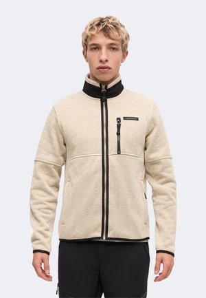 Jonge man met een beige gestructureerd zip-up jack met zwarte kraag en ritsdetails, staand tegen een eenvoudige lichte achtergrond.