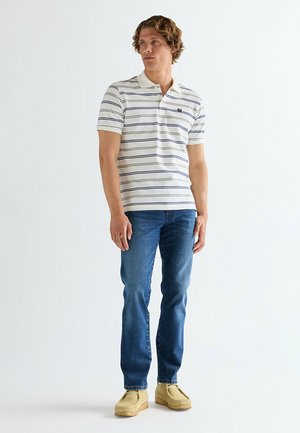 Młody mężczyzna stojący w białej koszulce polo z cienkimi granatowymi paskami, niebieskich dżinsach i beżowych zamszowych butach na jednolitym białym tle.