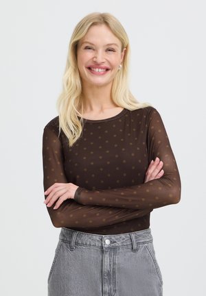 Femme blonde souriante, bras croisés, portant un haut marron transparent à manches longues à pois et un jean gris taille haute.
