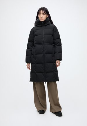 Woman wearing a long black padded coat with high collar, standing with hands relaxed by sides, wearing loose brown pants and black shoes.