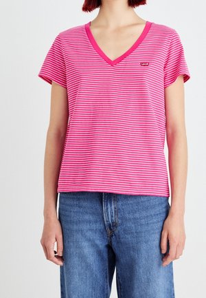 Femme portant un t-shirt à col en V rayé rose et blanc avec le logo Levi's et un jean bleu sur un fond uni.