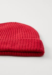 Bonnet tricoté rouge avec une texture côtelée et un revers replié, fabriqué en laine douce. Présente une trame lisse et uniforme sans motifs visibles.