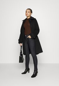 Abrigo largo negro sobre un suéter marrón de canalé, jeans oscuros y botines negros. Sosteniendo un pequeño bolso negro texturizado.