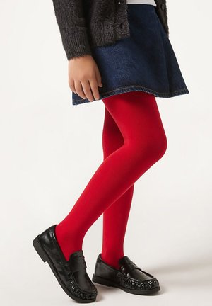 Enfant portant des collants rouge vif, une jupe bleu foncé, un pull gris et des mocassins noirs, debout avec une jambe croisée devant l'autre.