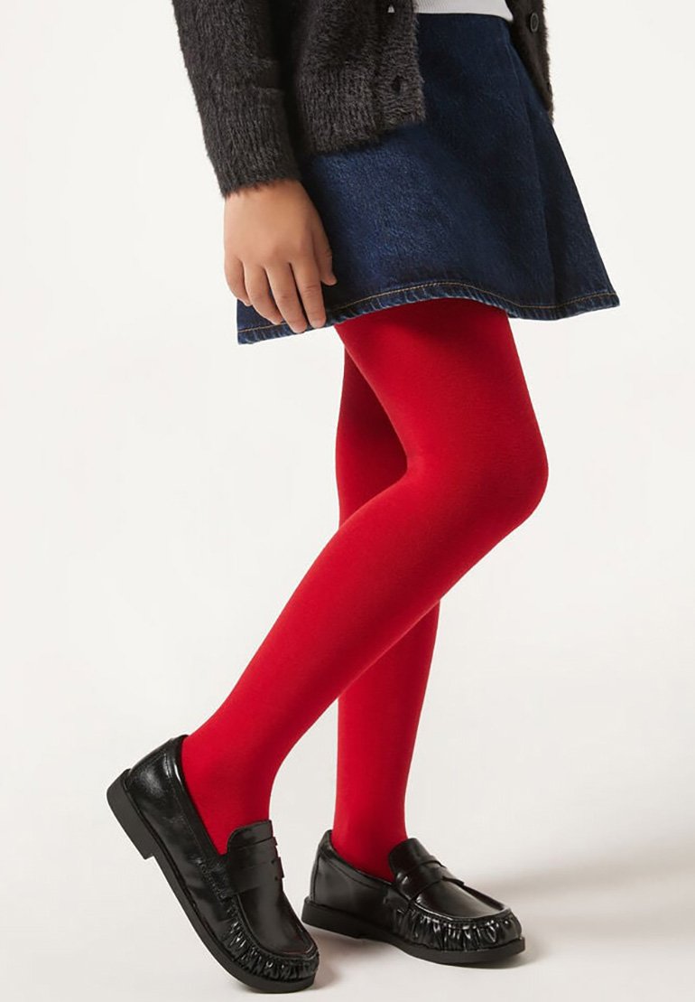 Enfant portant des collants rouge vif, une jupe bleu foncé, un pull gris et des mocassins noirs, debout avec une jambe croisée devant l'autre.