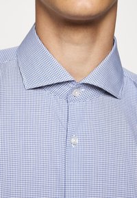 Gros plan d'une personne portant une chemise habillée à petits carreaux bleu et blanc avec un col pointu et deux boutons blancs fermés.