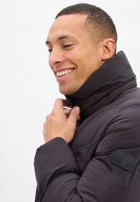 Zwarte pufferjas met hoge kraag, ritssluiting en logo-patch op de schouder. De gladde textuur en het gestikte ontwerp verhogen de warmte.