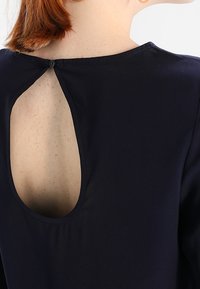 Námořnická blůza s otvoru na zádech se zapínáním na knoflík. Hladká tkanina s jemným leskem a bez viditelných vzorů či doplňků.