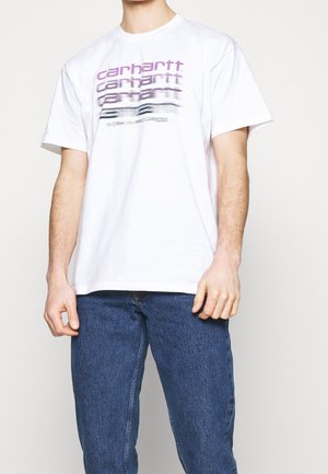 Homme portant un T-shirt blanc à manches courtes avec le texte délavé « Carhartt » en violet et noir, associé à un jean bleu sur fond uni.