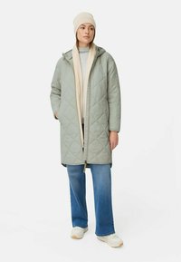 Kvinden bærer en lys grøn quiltet hættejakke, beige tørklæde og hue, grå sweater, blå jeans og hvide sneakers, stående mod en hvid baggrund.