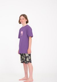 Camiseta de algodón morada con un gráfico de medusa, combinada con pantalones cortos negros que presentan patrones acuáticos coloridos. Descalzo sobre un fondo sencillo.